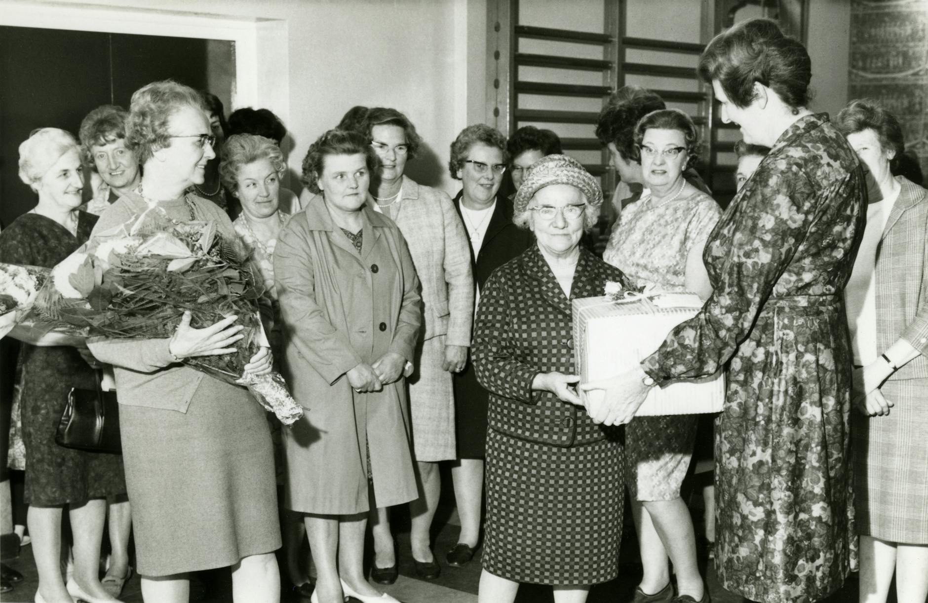 A group of elderly women in a vintage award ceremony receiving gifts and flowers.
